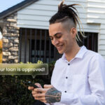 Young Kiwi man smiling at his phone after receiving a loan approval notification, standing outside a New Zealand home.