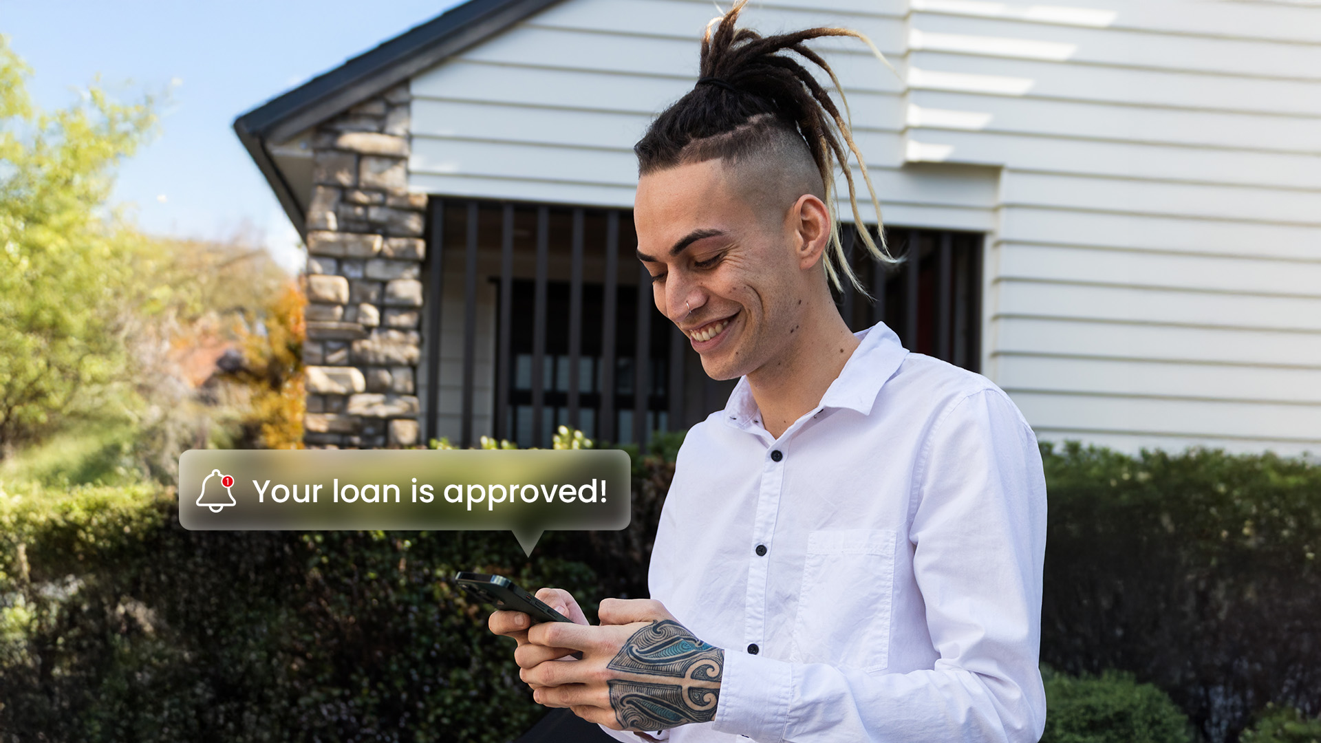 Young Kiwi man smiling at his phone after receiving a loan approval notification, standing outside a New Zealand home.